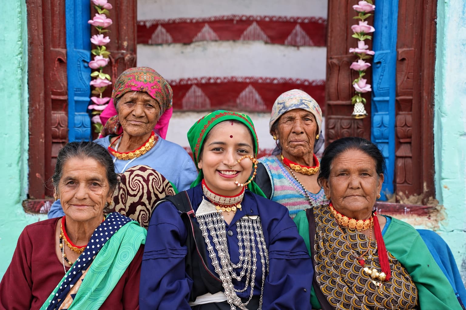 Five women wearing traditional Pahadi jewellery in front of a carved blue and red doorway